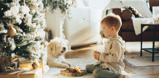 Are You Worried Your Little Elves Are Getting S”Elf”ish This Holiday Season? A toddler sitting by a Christmas tree.
