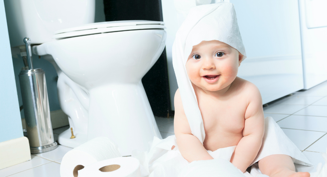 A baby wrapped in toilet paper sitting next to a toilet.