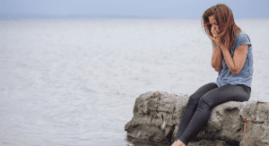 Woman holding her face sitting on the rocks by the water.