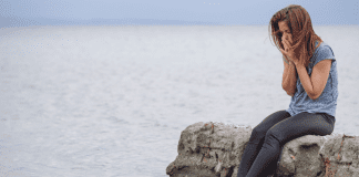 Woman holding her face sitting on the rocks by the water.