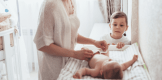 A woman changing a baby's diaper while her toddler watches.