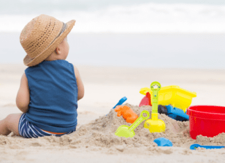 A baby boy sitting on the beach next to his sand toys.