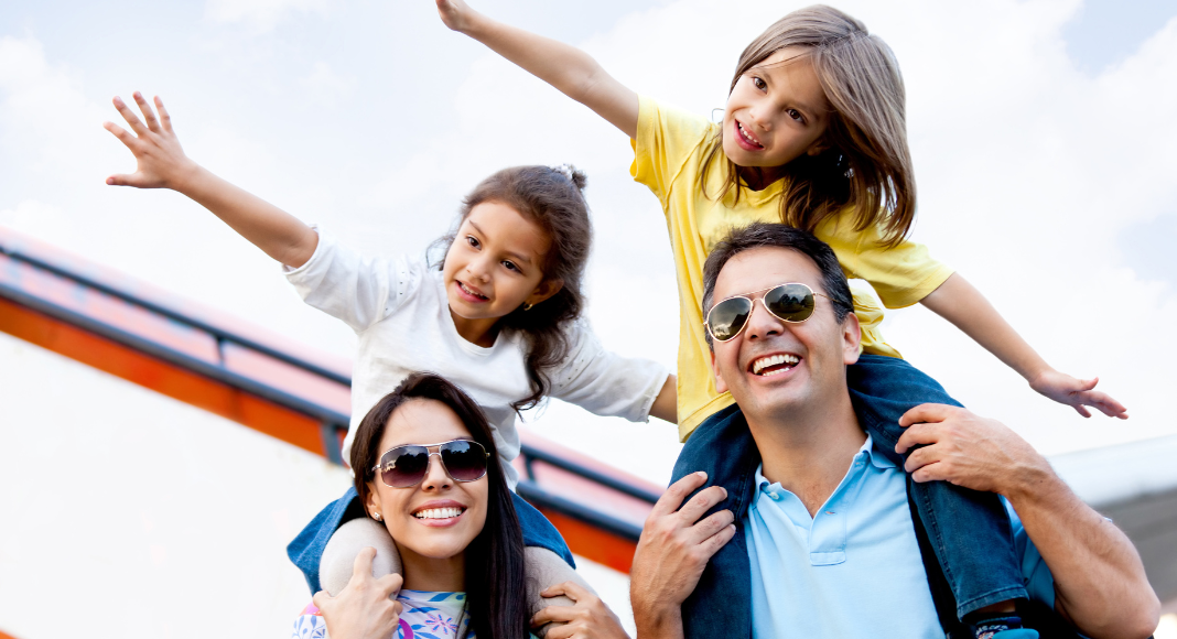 A family of four. The kids are sitting on the parent's shoulders with their arms out. 