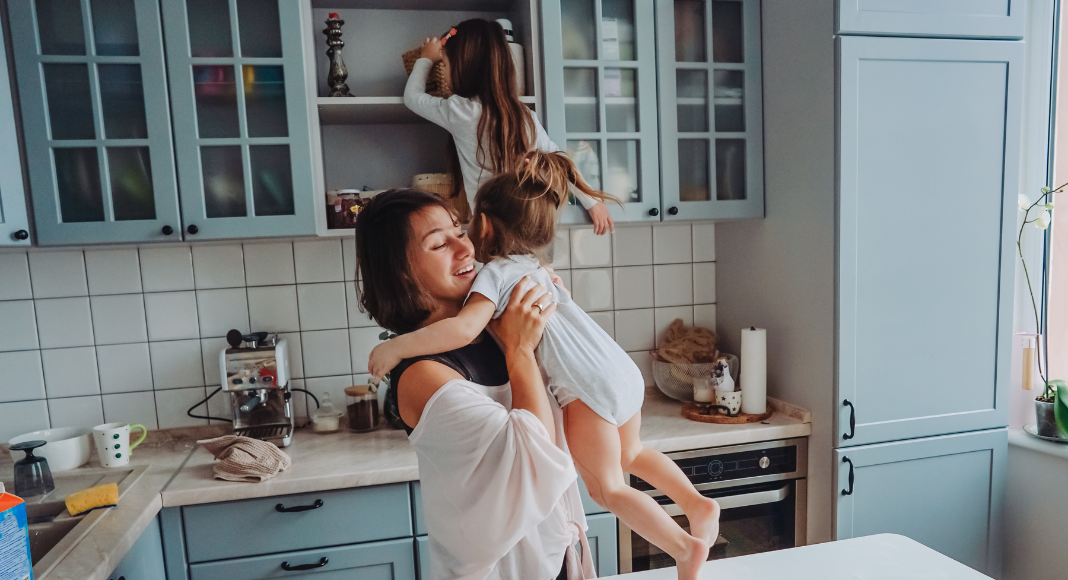 A mom in the kitchen with her daughters climbing on the counters.