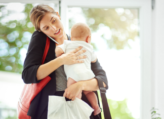 A mom on the phone holding her baby and lots of bags.