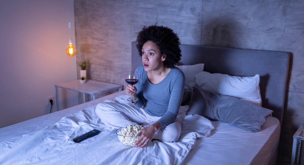 A woman alone watching TV at night drinking wine and eating popcorn.