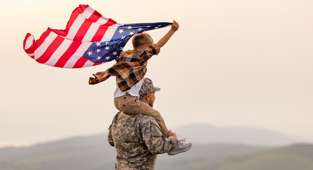 A veteran holding his son on his shoulders as the boy holds the American flag. 